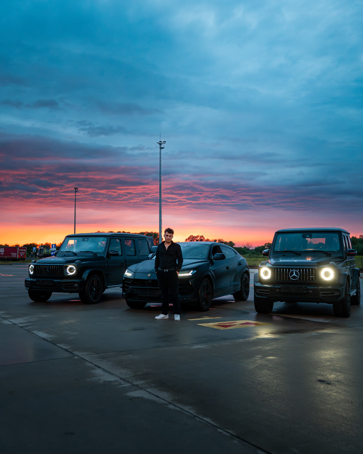 Gerox przed czarnym Lamborghini Urus, po bokach dwa czarne Mercedesy G 63, zachód słońca